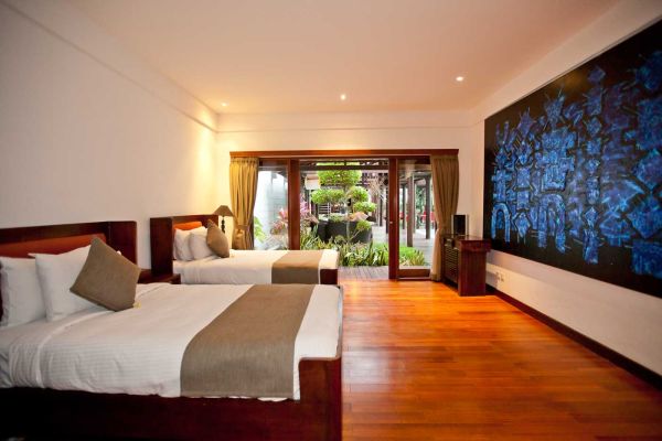 Spacious Balinese villa twin bedroom with wooden beds, neutral linens, large blue abstract wall art, and tropical garden view through open sliding doors.