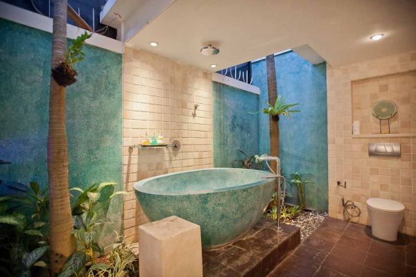 Luxurious tropical outdoor bathroom with large turquoise freestanding bathtub, lush plants, palm trees, and stone features. - Bali Villas