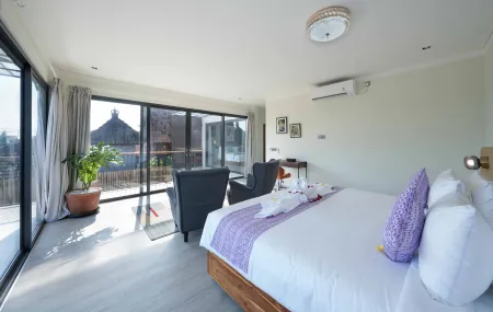 Spacious Bali villa bedroom with king bed, purple accents, cozy armchairs, and tropical balcony view through floor-to-ceiling glass doors.