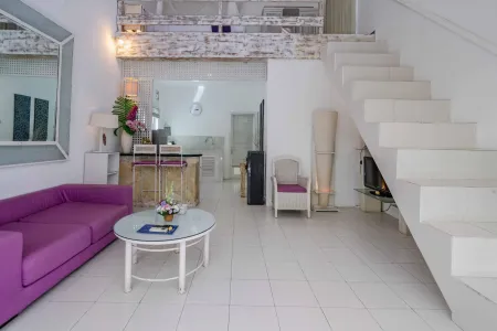 Modern loft apartment interior with purple sofa, glass coffee table, wooden kitchenette, potted flowers, and white staircase. - Bali Villas