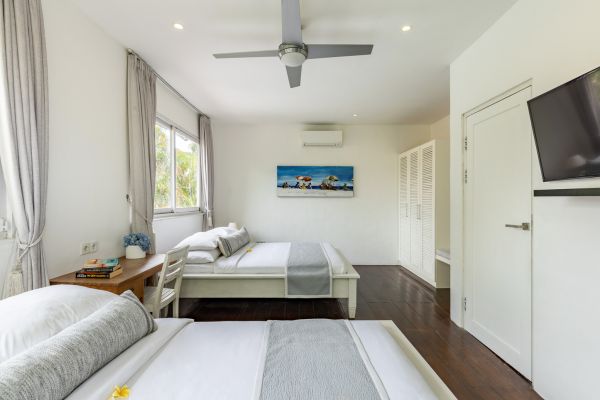 Spacious Bali villa bedroom with king bed, white linens, ceiling fan, AC, louvered wardrobe, and tropical garden views through sheer-curtained windows.