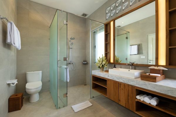 Modern bathroom interior with glass shower enclosure, wooden vanity, white sink, toilet, and fresh flowers. - Bali Villas