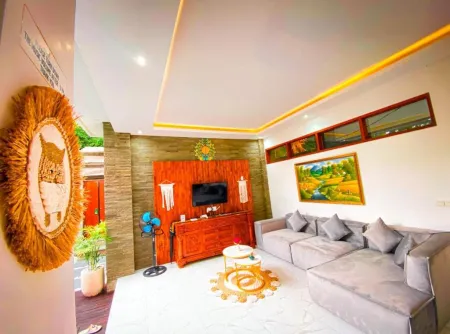 Cozy modern Balinese living room with gray L-shaped sofa, wooden TV cabinet, woven owl wall hanging, landscape artwork, and tropical decor. - Bali Villas