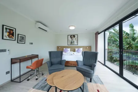 Modern luxury hotel bedroom with king-sized bed, gray armchairs, wooden desk, and sliding glass doors opening to a tropical balcony.