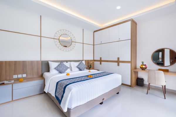 Spacious modern Bali bedroom with king bed, blue batik runner, tropical flowers, wooden wardrobe, and round sunburst mirror.