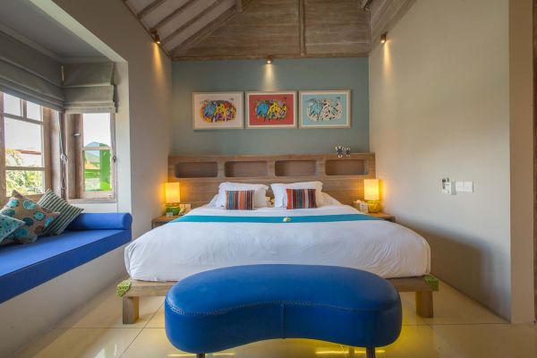 Cozy Balinese bedroom interior with wooden bed, colorful abstract wall art, turquoise accents, and tropical garden view through shuttered windows.