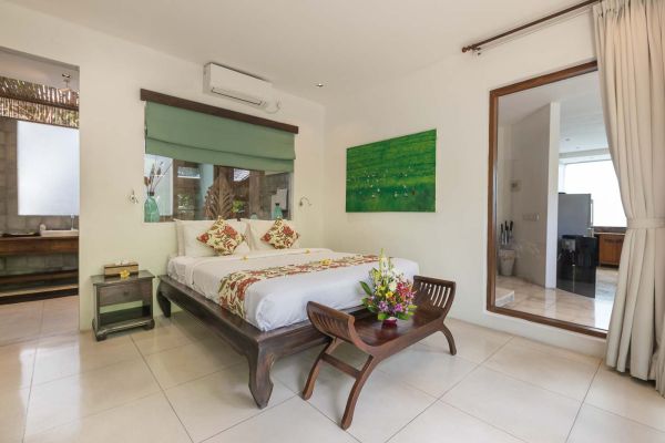 Luxurious Bali-inspired bedroom with a king bed, floral accents, green abstract painting, and open door to en-suite bathroom.