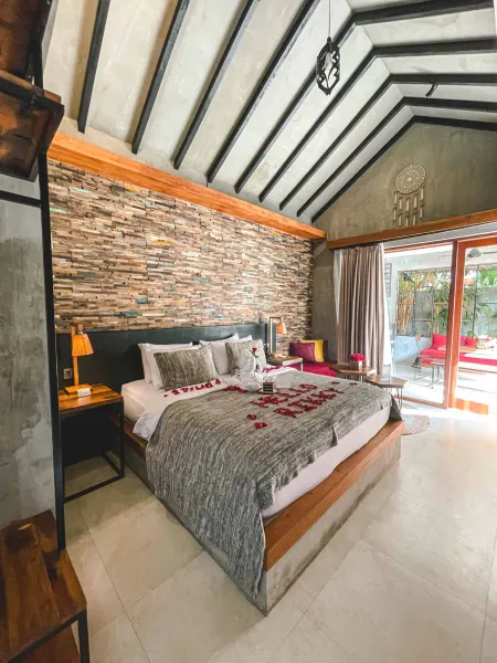 Romantic Balinese villa bedroom with king bed scattered in red rose petals spelling "LOVE", wooden beamed ceiling, and sliding doors opening to a lush garden.