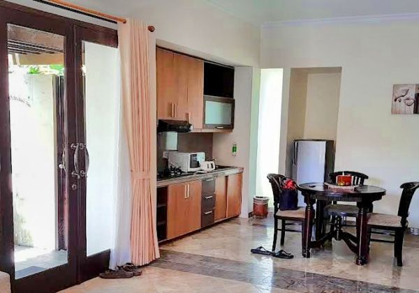 Cozy tropical kitchenette with wooden cabinets, stove, microwave, fridge, and small dining table in a light-filled room with open wooden doors and pink curtains. - Bali Villas