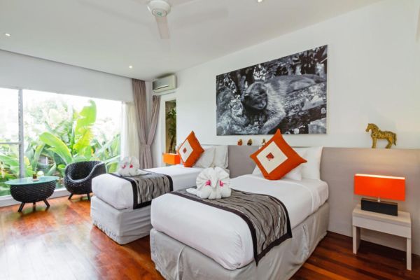 Spacious tropical bedroom in a Bali villa with two white twin beds, orange accents, large black-and-white wall art, and lush garden view through sliding doors.