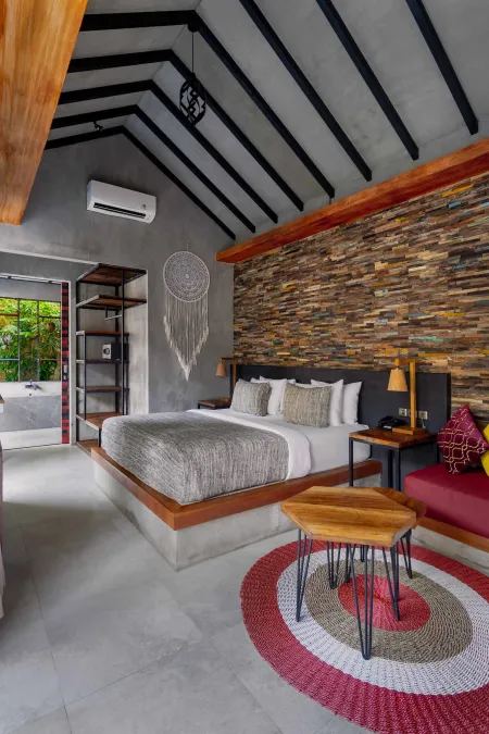 Luxurious tropical bedroom with reclaimed wood walls, king bed on concrete platform, dreamcatcher decor, wooden side tables, and red circular rug.