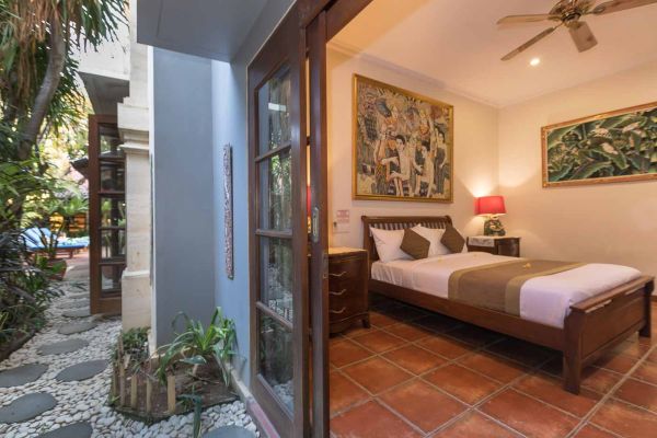 Cozy Balinese villa bedroom with wooden bed, traditional artwork, ceiling fan, and open doors to lush tropical garden and pool.