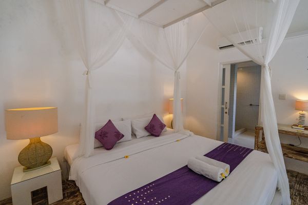 Elegant Balinese bedroom with white canopy bed, purple pillows and runner, bedside lamps, and open bathroom door.