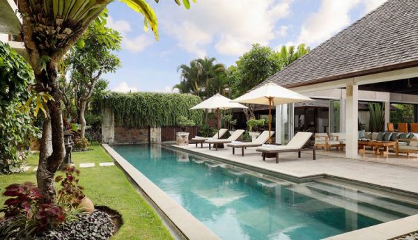 Serene infinity pool at a luxurious Balinese villa, with lounge chairs, white umbrellas, tropical palms, and lush gardens under a partly cloudy sky. - Bali Villas