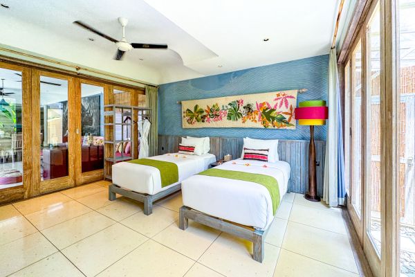 Cozy tropical resort bedroom with two twin beds, blue floral wall art, wooden sliding doors to garden, and ceiling fan.
