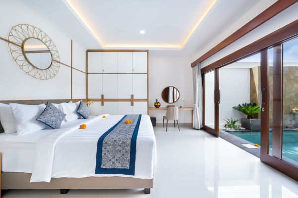 Spacious Balinese villa bedroom with king bed, blue runner, vanity mirror, and sliding doors opening to private pool.