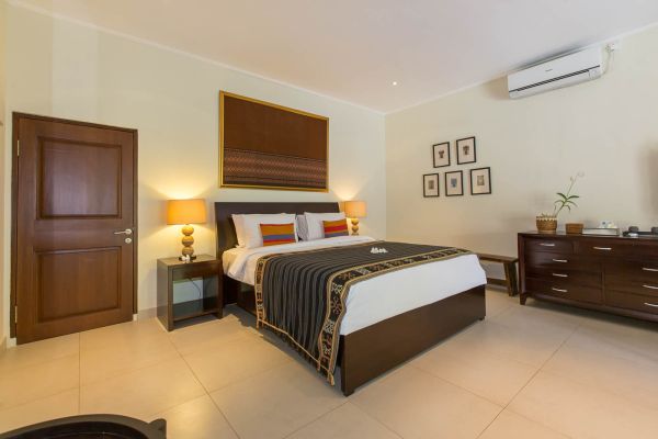 Elegant Balinese-style bedroom with dark wooden king bed, orange accent pillows, bedside lamps, wall art, and potted plant on dresser.