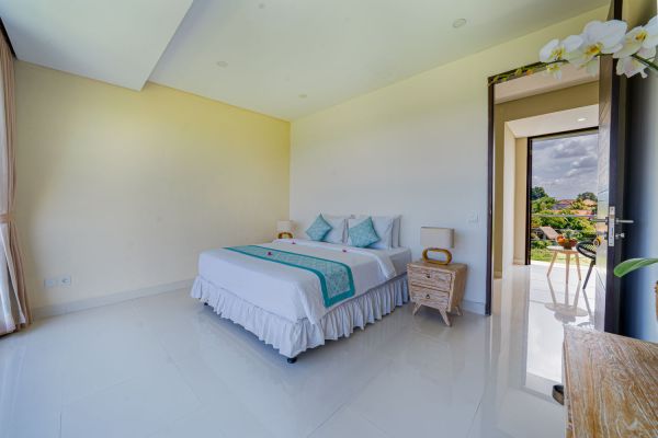Spacious Balinese bedroom with white bed, teal accents, wooden furniture, orchid plant, and open door to tropical outdoor view.