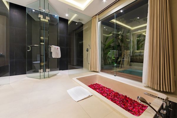 Luxurious modern bathroom with freestanding bathtub filled with red rose petals, glass shower enclosure, and large windows overlooking tropical greenery. - Bali Villas