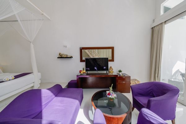 Luxurious Balinese villa bedroom with white linens on a canopy bed, purple upholstered sofas, wooden TV cabinet, and sliding glass doors to outdoor terrace.