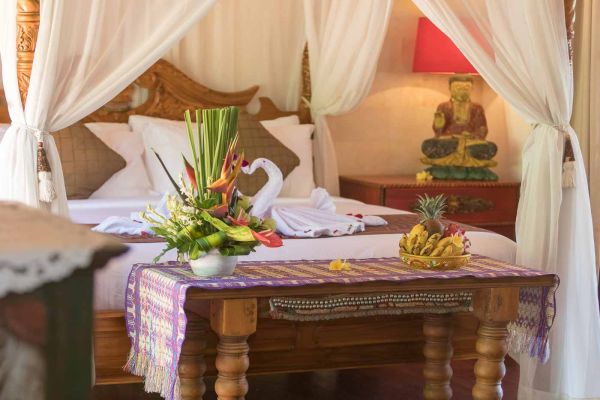 Luxurious tropical bedroom with canopy bed, white linens, vibrant flower arrangement, towel swans, and bedside table with Buddha statue and fresh pineapple.