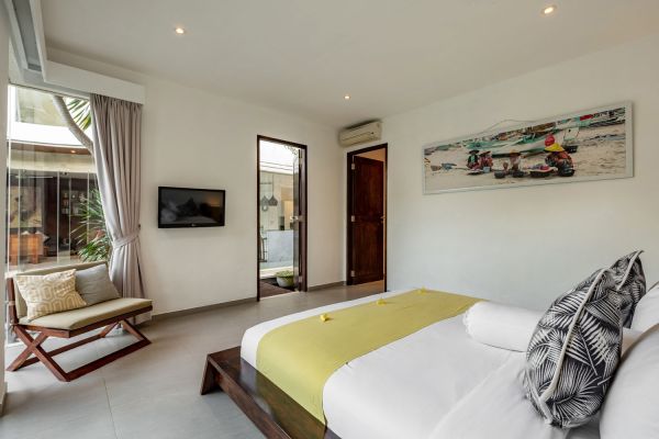 Spacious modern Bali villa bedroom with king bed, yellow bedding, mounted TV, and sliding doors to tropical courtyard.