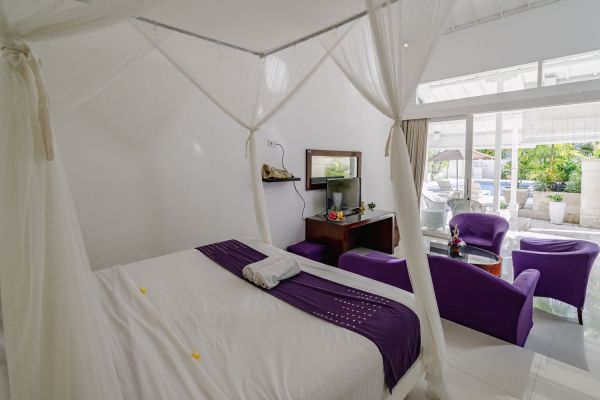 Elegant Balinese-style bedroom with white canopy bed, purple accents, and tropical pool view through sliding doors.