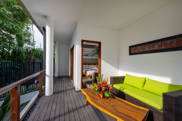 Luxurious tropical balcony with green rattan sofa, wooden coffee table topped with vibrant flowers, and sliding doors to a serene bedroom.
