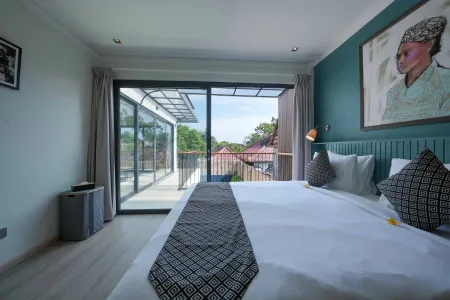 Spacious modern Bali villa bedroom with sliding glass doors to balcony overlooking tropical garden, king bed with frangipani flower, and vibrant wall art of a woman in headwrap.