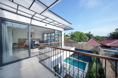 Modern tropical villa balcony with glass doors to bedroom, overlooking private turquoise pool and lush greenery under blue sky.