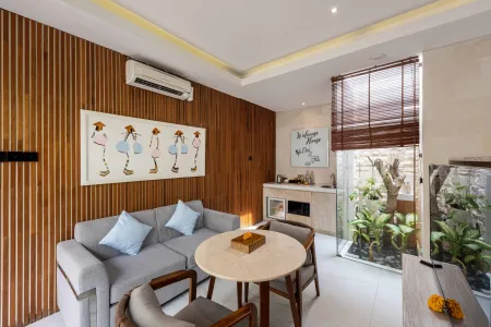 Cozy Balinese villa lounge with wooden paneled walls, gray sofa and chairs around a marble table, abstract wall art, and lush garden window view. - Bali Villas