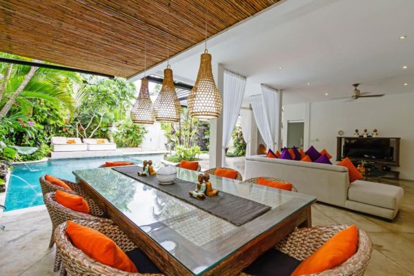 Luxurious open-air tropical villa with bamboo ceiling, hanging rattan lamps, glass dining table in orange chairs, white sofa with vibrant cushions, and infinity pool amid lush greenery. - Bali Villas