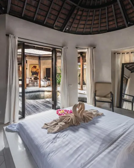 Serene Balinese villa bedroom with thatched roof, white bed featuring swan-shaped towel and hibiscus flowers, and glass doors opening to a private pool.