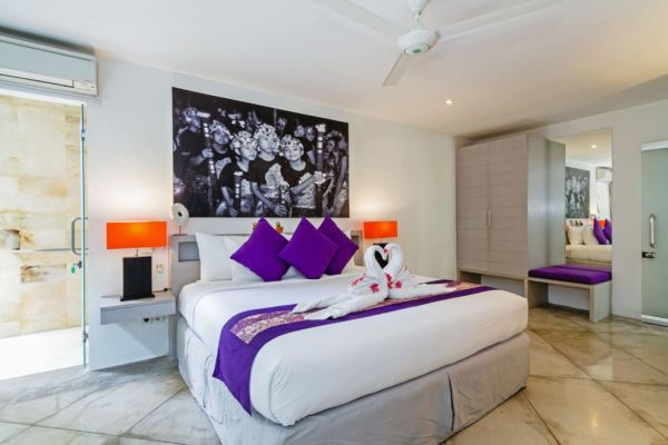 Luxurious Bali-style hotel bedroom with white king bed, purple bedding, swan towels, orange lamps, and black-and-white wall mural. - Bali Villas