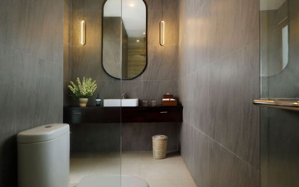 Modern minimalist bathroom featuring gray stone tiles, floating vanity with sink and oval mirror, toilet, walk-in glass shower, and potted plant. - Bali Villas