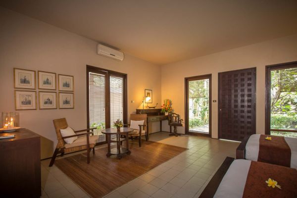 Spacious Balinese villa bedroom with king bed topped by frangipani flowers, wooden armchairs, desk, and tropical garden views through sliding doors.