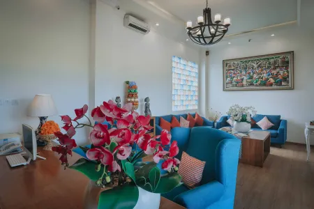 Modern hotel lounge with blue upholstered sofas, vibrant pink orchids on a wooden desk, colorful mosaic window, and a large tropical wall painting under chandelier lighting. - Bali Villas
