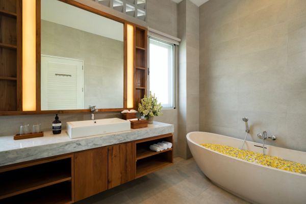 Luxury spa bathroom with wooden vanity, white freestanding bathtub filled with yellow frangipani petals, and neutral tones. - Bali Villas