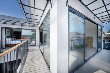 Modern tropical balcony with wooden deck, metal railing, and large sliding glass doors to white-walled bedroom, overlooking lush greenery.