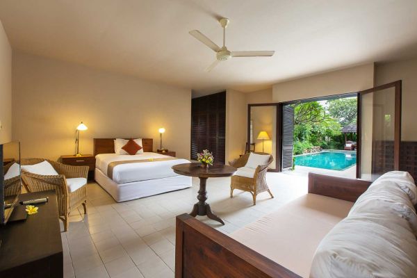 Spacious tropical bedroom in a luxury villa, featuring a king bed, rattan furniture, and French doors opening to a private pool amid lush greenery.