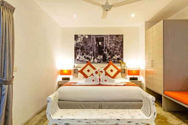 Elegant tropical bedroom with white king bed, red linens, swan towels, bedside lamps, ceiling fan, and large black-and-white cityscape artwork above.