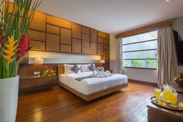 Luxurious Balinese villa bedroom with king-sized bed, tropical orchids, fresh orange juice tray, and garden view through large wooden-framed window.