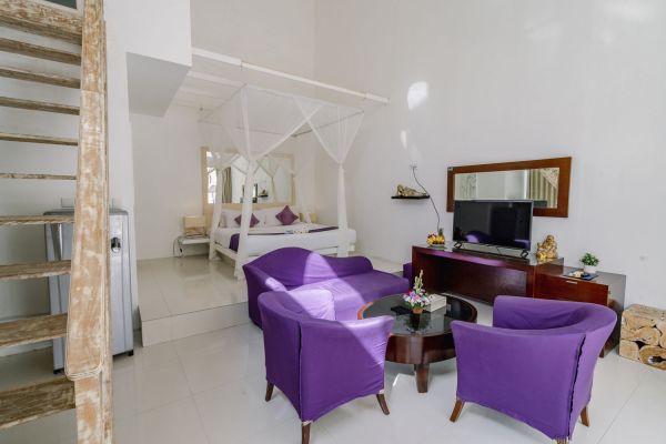 Bright Balinese villa bedroom with white canopy bed, purple armchairs around a coffee table, wall-mounted TV, and wooden loft stairs.