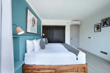 Modern tropical bedroom with wooden king bed, crisp white linens, teal accent wall, framed artwork, and ambient lighting.