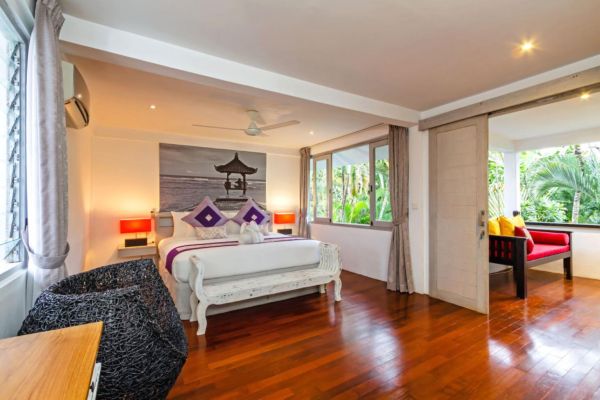 Spacious Bali villa bedroom with white wooden bed, purple accents, ceiling fan, and lush garden views through sliding doors.