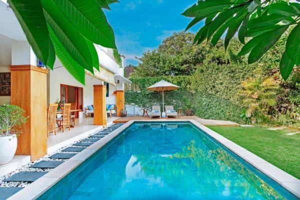 Modern tropical backyard with a long blue rectangular pool, wooden patio furniture, lounge chairs, and lush green plants under a clear sky. - Bali Villas