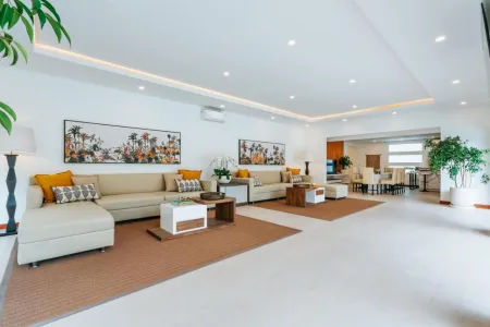Modern spacious living room with beige L-shaped sofas, wooden coffee tables, palm tree wall art, orange accents, and potted plants in a bright white interior. - Bali Villas