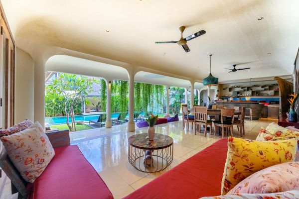 Luxurious open-plan Bali villa living room with red sofa, wooden dining area, and poolside garden views. - Bali Villas