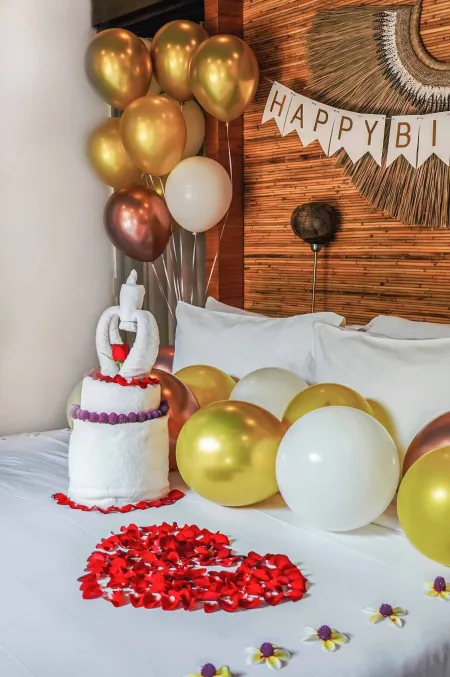Romantic birthday bedroom surprise: towel cake with red ribbon on bed surrounded by gold, rose gold, and white balloons, red rose petal heart, and woven wall decor with "Happy Birthday" banner.
