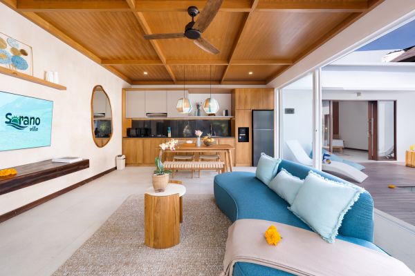 Spacious Balinese villa living room with wooden beamed ceiling, open kitchen, blue sofa, and infinity pool view. - Bali Villas